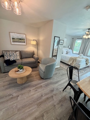 Ein modernes Wohnzimmer mit einem grauen Sofa, einem runden Couchtisch mit Pflanze, einem grauen Sessel und einem Essbereich mit Holztisch und Stühlen.