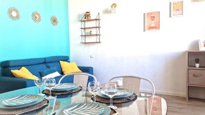 Un'area pranzo con un tavolo in vetro apparecchiato per quattro persone, un divano blu con cuscini gialli e una mensola in legno con oggetti decorativi.
