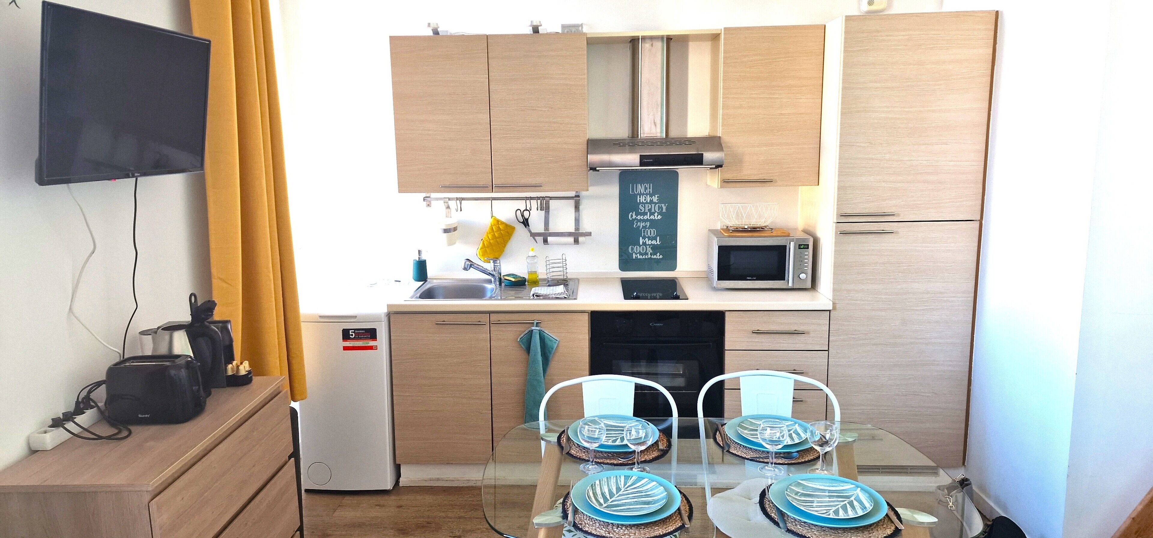 A compact kitchen with a microwave, toaster, and small refrigerator. There is a dining table with chairs and a TV mounted on the wall.