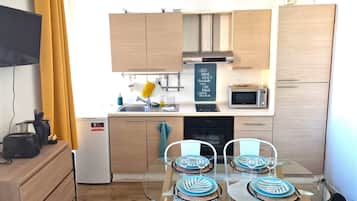 A compact kitchen with a microwave, toaster, and small refrigerator. There is a dining table with chairs and a TV mounted on the wall.