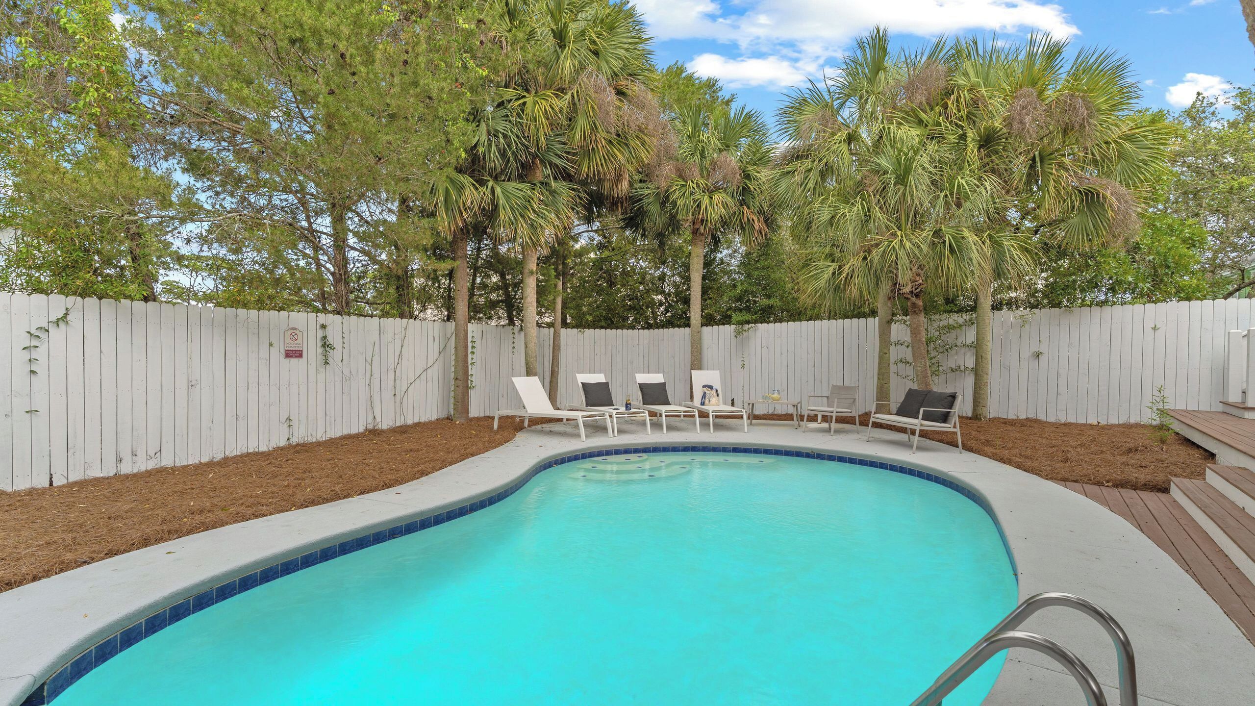 A swimming pool with lounge chairs, surrounded by a white fence and trees.