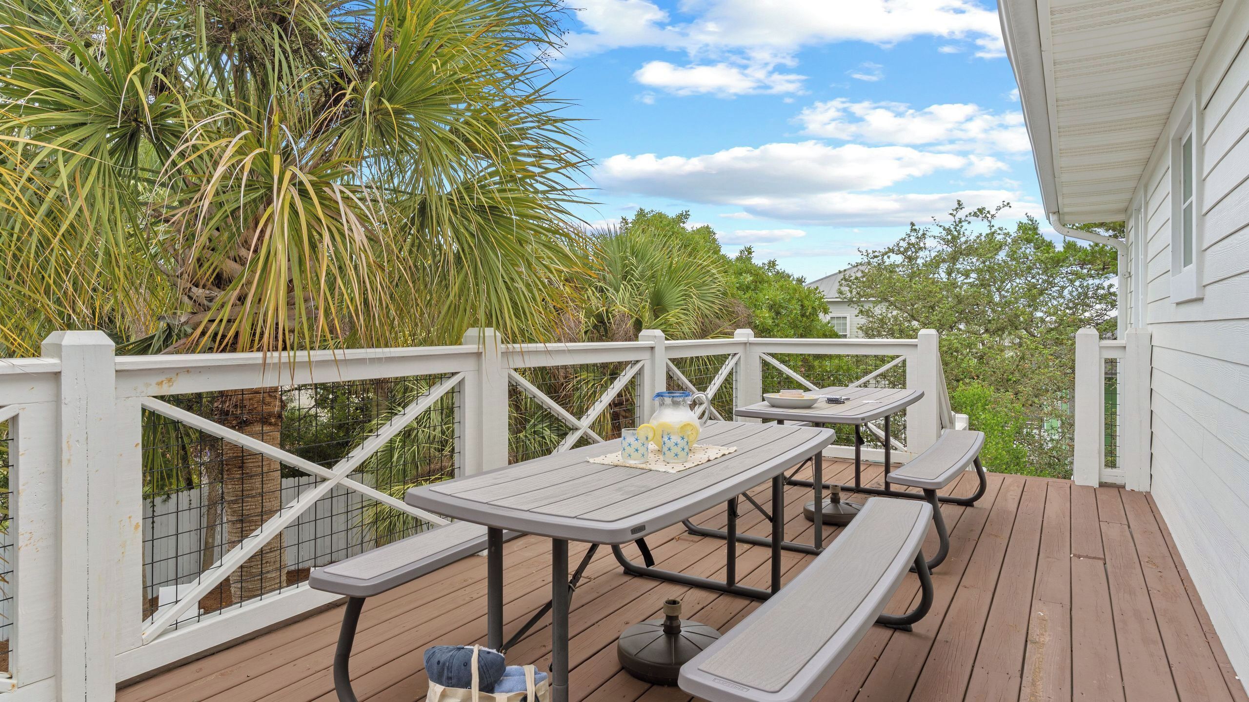Une terrasse en bois, agrémentée de tables de pique-nique et de bancs, entourée d’une clôture blanche et d’une végétation luxuriante.