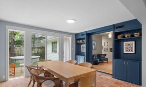 Een eetruimte met een houten tafel en rieten stoelen, een blauwe accentmuur met planken, en uitzicht op de achtertuin door glazen deuren.