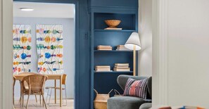 Een kamer met een eettafel en stoelen, een boekenplank met boeken en een lamp, en een bank met een gestreept kussen.