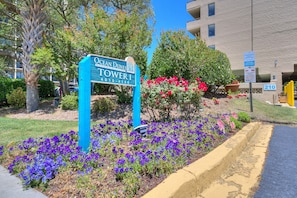 Schild für Ocean Dunes Tower I, ein Gebäude, das von gepflegten Gärten mit violetten Blumen, einer Palme und blauem Himmel umgeben ist.