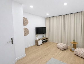 A modern living room with a flat-screen TV, wooden flooring, a sofa, a small side table, and a curtain.