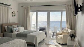 Een moderne slaapkamer met twee bedden, een plafondventilator, een grote schuifglazen deur naar een balkon en uitzicht op zee.