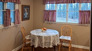 Une salle à manger avec une table ronde, deux chaises, un saladier de fruits et des rideaux à carreaux.