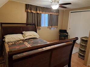 A bedroom with a wooden bed, two pillows, a window with checkered curtains, a ceiling fan, and a storage unit.
