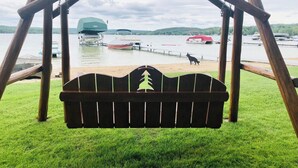 Ein hölzernes Schaukelgerüst mit einer Bank davor, mit Blick auf einen See mit Booten und einem Hund am Ufer.