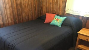 Ein Bett mit dunkelblauer Bettdecke und zwei Kissen, eines rot und eines mit Vogelmotiv, in einem Zimmer mit Holzwänden und einem Nachttisch aus Holz.