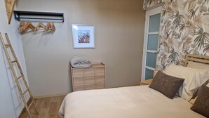 Ein Schlafzimmer mit einem Bett, einem hölzernen Schminktisch, einem gerahmten Bild, einem Kleiderständer und einem Fenster mit einem blattbedruckten Vorhang.