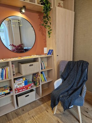 Habitació amb mirall, prestatges amb llibres i jocs, una cadira amb una manta i una planta.