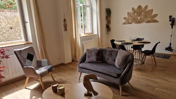 A modern living room with a grey sofa, a dining table with chairs, and a large window with curtains.