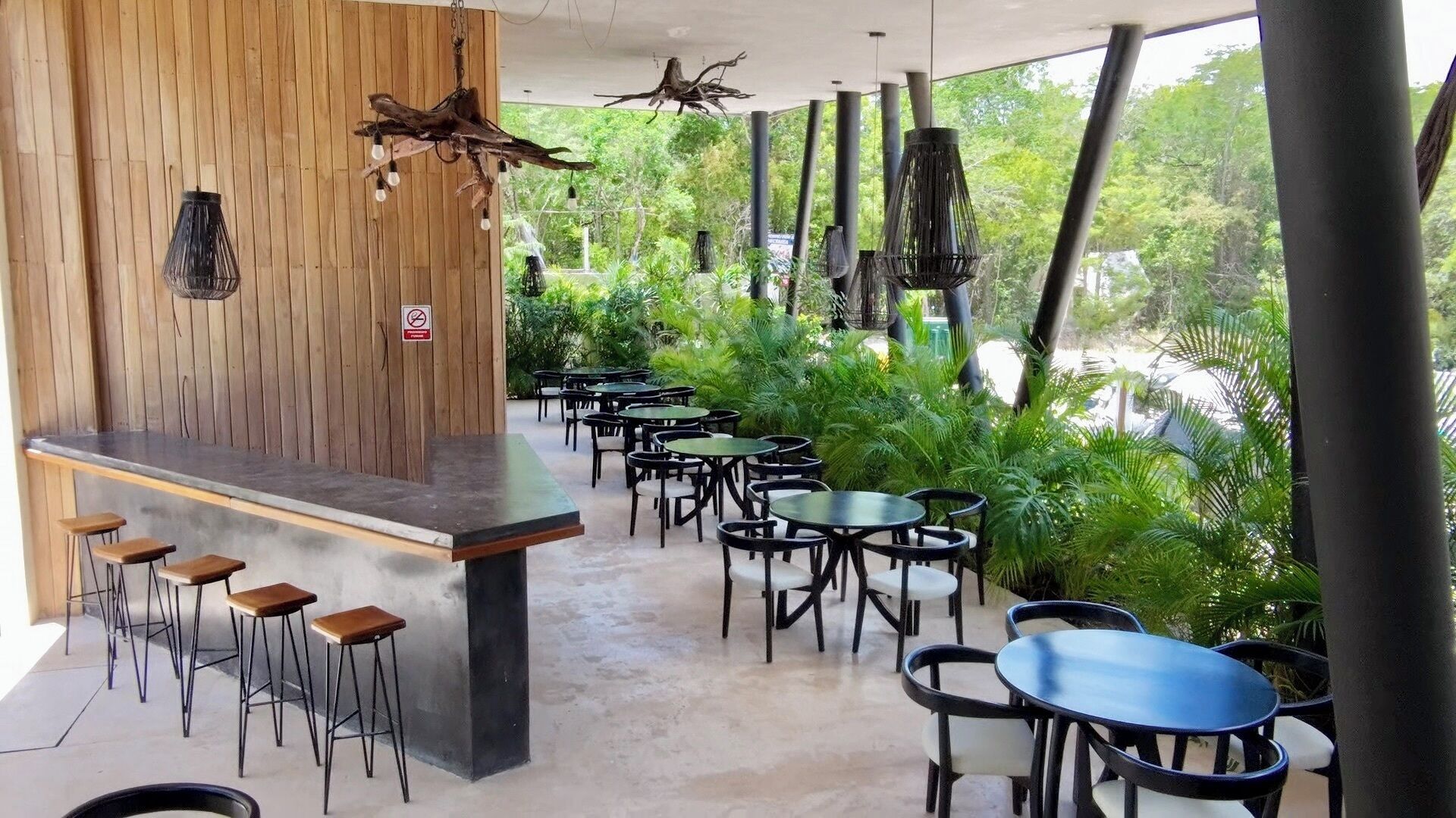 An outdoor cafe with wooden walls, a bar area with stools, and tables surrounded by greenery.