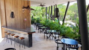 Um café ao ar livre com paredes de madeira, área de bar com banquetas e mesas rodeadas por vegetação.