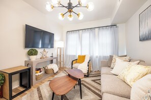 Ein modernes Wohnzimmer mit Flachbildfernseher, einer Couch, einem Couchtisch, einem Sessel und einer Deckenleuchte.