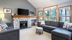 A living room with a stone fireplace, a flat-screen TV, a grey sofa, and large windows. - Steller's Jay Chalet - private hot tub, ski in/out, 2 bedroom, 2 bath townhome (Whistler)