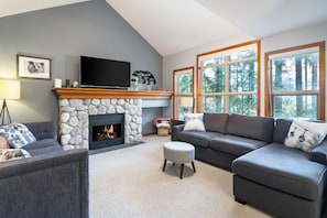 Una sala de estar con chimenea de piedra, televisor de pantalla plana, sofá gris y ventanas amplias.