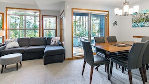 A modern living room with a sectional sofa, a dining table with chairs, large windows, and a view of greenery. - Steller's Jay Chalet - private hot tub, ski in/out, 2 bedroom, 2 bath townhome (Whistler)