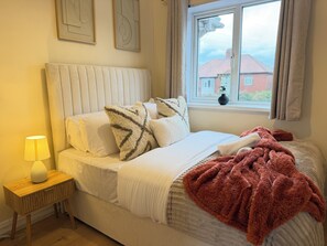 Ein Bett mit Kopfteil, Kissen, Decke, einem Nachttisch mit Lampe und einem Fenster mit Blick auf Gebäude.