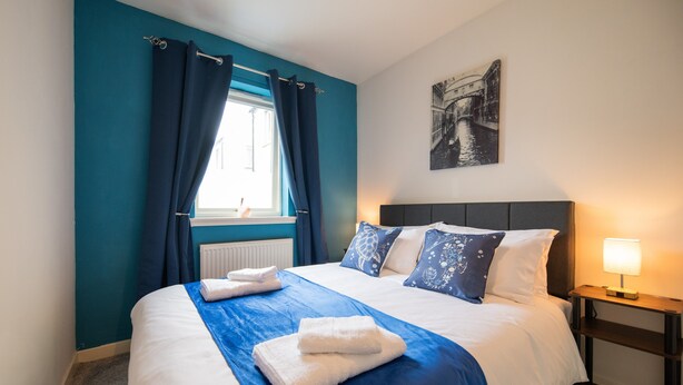 A neatly made bed with white and blue bedding, a window with blue curtains, a bedside table with a lamp, and a wall-mounted picture.