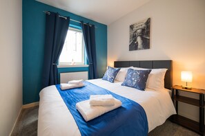 A neatly made bed with white and blue bedding, a window with blue curtains, a bedside table with a lamp, and a wall-mounted picture. - Surfer 2Bed GF Flat Central Thurso, Parking, Full Fibre, Workspace, Pet friendly (Thurso)