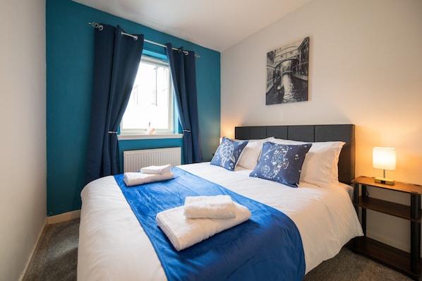 A neatly made bed with white and blue bedding, a window with blue curtains, a bedside table with a lamp, and a wall-mounted picture.