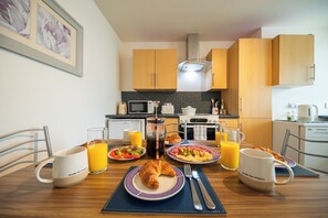 A breakfast table with croissants, plates of food, and glasses of orange juice in a modern kitchen with wooden cabinets and a microwave. - Surfer 2Bed GF Flat Central Thurso, Parking, Full Fibre, Workspace, Pet friendly (Thurso)