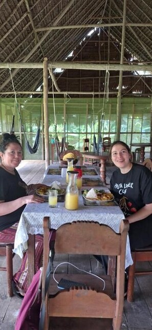 Duas pessoas sentadas à mesa com toalha branca, uma rede e uma estrutura de telhado de palha.