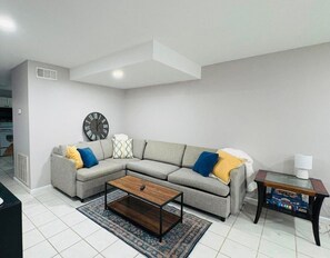 Ein Wohnzimmer mit grauem Sofa, einem Holztisch, einer Wanduhr und einem kleinen Tisch mit Lampe und Büchern.
