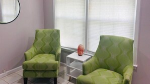 Una habitación con dos sillones verdes con estampado, una mesita blanca con un objeto decorativo y un ventanal con persianas.