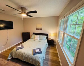 Ein Schlafzimmer mit Bett, Deckenventilator, am Wand montiertem Fernseher, Fenster mit Blick auf Grünflächen und Nachttisch mit Lampe.