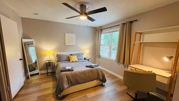 Un dormitorio con cama, lámparas de noche, escritorio, silla, ventilador de techo y ventana con cortinas.