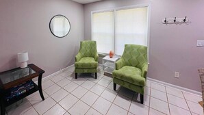 Una habitación con dos sillones verdes, una mesita, un espejo redondo y un ventanal con persianas.