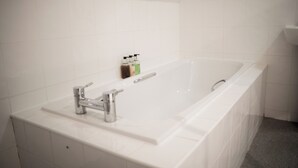 A modern bathroom with a white bathtub, chrome fixtures, and a small shelf with toiletries. - Family King Room with Attached Single Room (Bude)