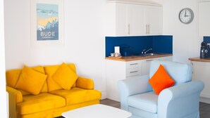 Una habitación con un sofá amarillo, un sillón azul claro, una mesa de centro blanca, una zona de cocina con armarios, un lavabo y un reloj en la pared.
