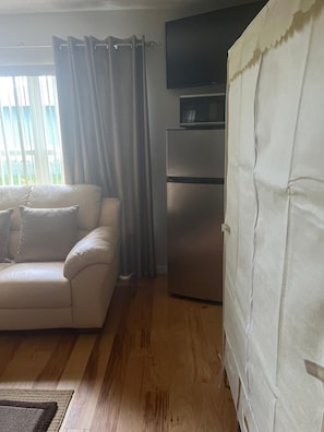 Una sala de estar con un sofá, un televisor de pantalla plana en la pared, un refrigerador y una ventana con cortinas.