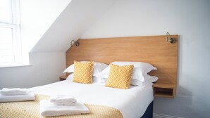 Habitación de hotel con cabecera de madera, dos almohadas, una toalla blanca doblada con cuidado y una colcha amarilla estampada.