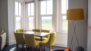 Een kamer met een eettafel en stoelen, een bank, een lamp en grote ramen.
