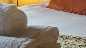 Una cama bien tendida con sábanas blancas, un almohadón marrón y una toalla blanca enrollada sobre una colcha texturizada.