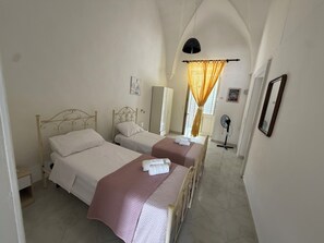 A bedroom with two beds, a standing fan, and a window with curtains.