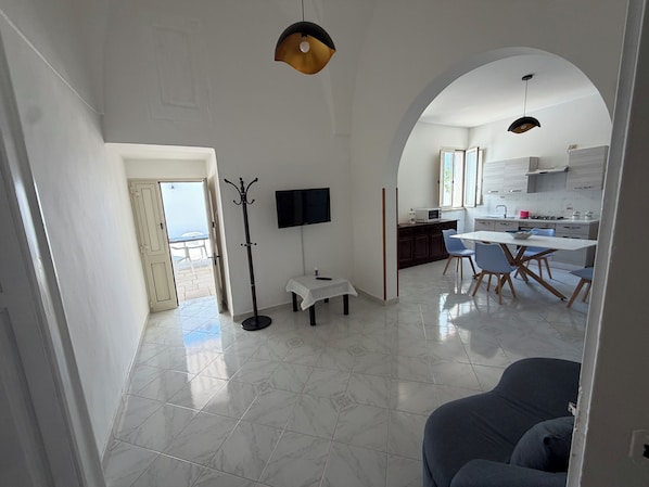 Una sala moderna con un sofá, un comedor con sillas, un perchero, un televisor de pantalla plana y una cocina al fondo.