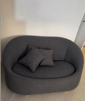 A dark grey sofa with two cushions, positioned against a plain wall with a tiled floor.