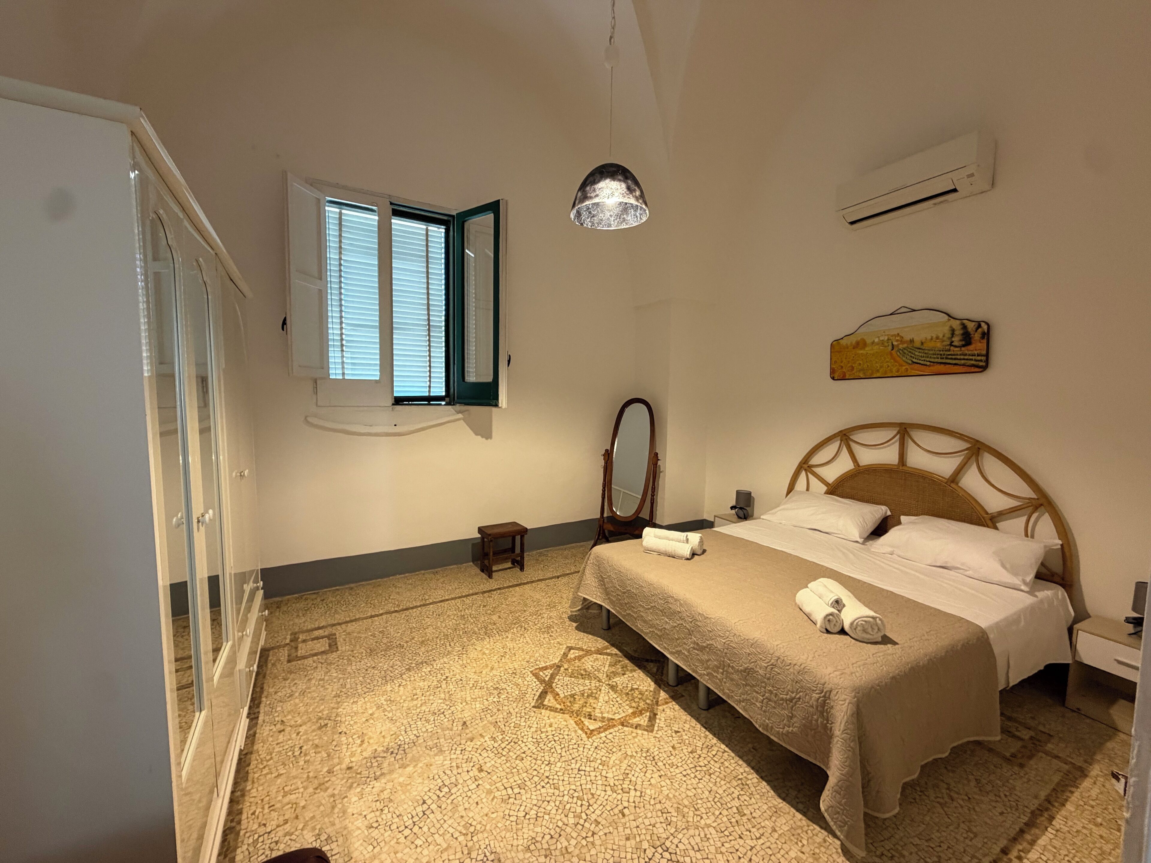 A bedroom with a bed, a bedside table, a mirror, a window with blinds, and a wall-mounted air conditioner.