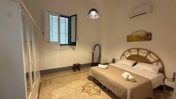 A bedroom with a bed, a bedside table, a mirror, a window with blinds, and a wall-mounted air conditioner.