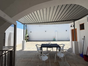 Un patio al aire libre con mesa y sillas, una parrilla y una pared con una puerta y ventanas.