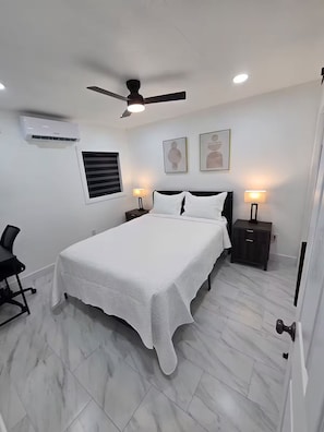 Ein Schlafzimmer mit Bett, Schreibtisch, Stuhl, Deckenventilator, Wandaircondition und gerahmten Kunstwerken.