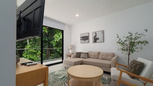 Una sala de estar moderna con un sofá, una mesa de centro redonda, un televisor sobre un mueble bajo y un ventanal con vistas a zonas verdes.