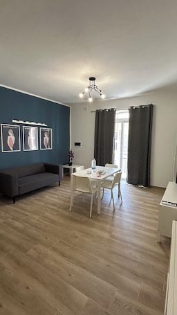 A modern living room with a sofa, dining table, and chairs, a ceiling light fixture, and a window with curtains.. Comfortable Apartment in Xlendi • Walk to the Sea